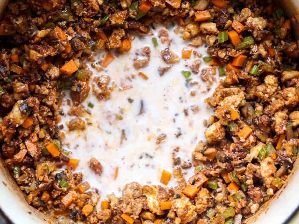Close-up of a pan with diced carrots, celery, onions, and crumbled tofu cooked around the edges, leaving a pool of non-dairy milk in the center being incorporated into the mixture.